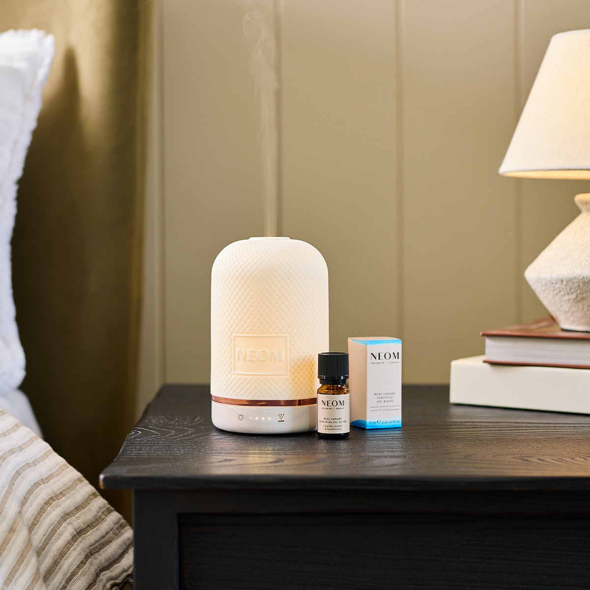 NEOM Wellbeing Pod releasing mist beside its Real Luxury essential oil blend on a bedside table