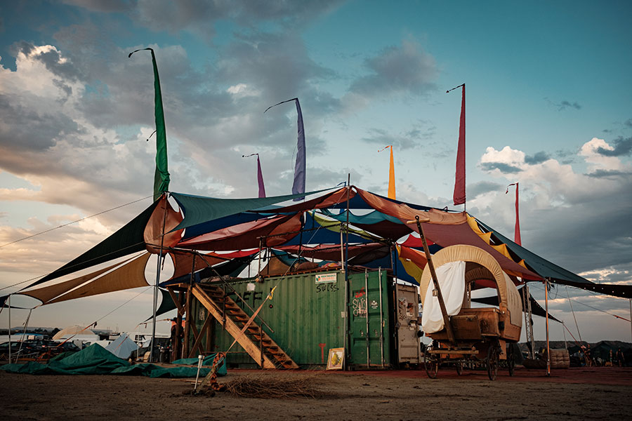 Colourful shade sails and tall flags above a green container and covered wagon at Nowhere