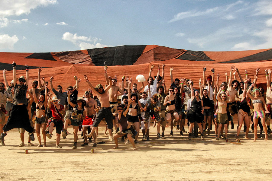 Large group of Nowhere participants raising their arms beneath an orange shade canopy