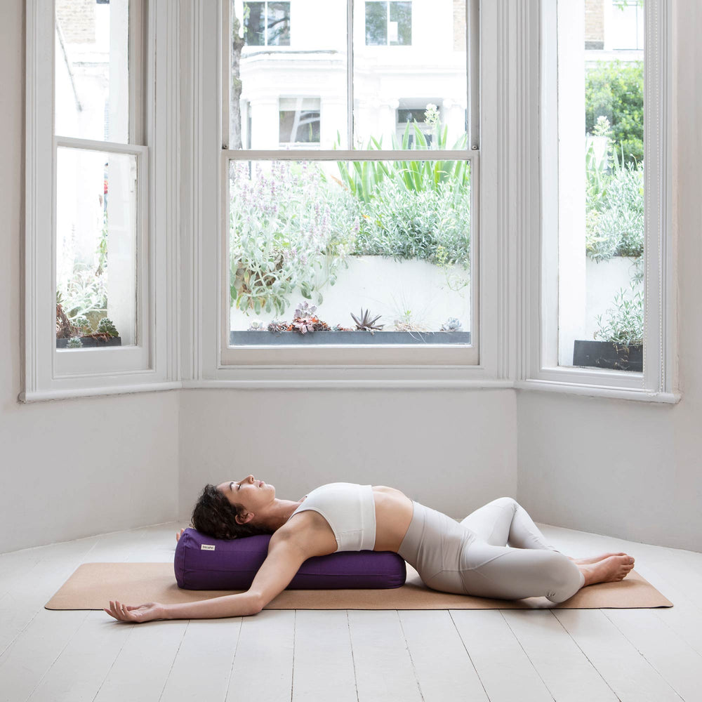 Woman reclining on a purple basaho yoga bolster beside a bright window